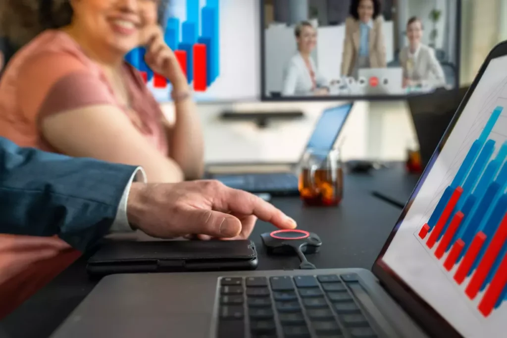 Hybrides Meeting mit Clickshare CX-50 der 2. Generation. Auf dem Tisch ist das Gerät und im Hintergrund erkennt man die Teilnehmer, die digital dazugeschalten sind.